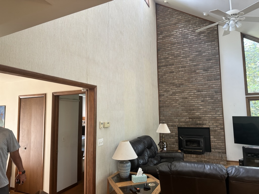 Original great room with dated grasscloth wallpaper, dark brick fireplace, and heavy wood trim